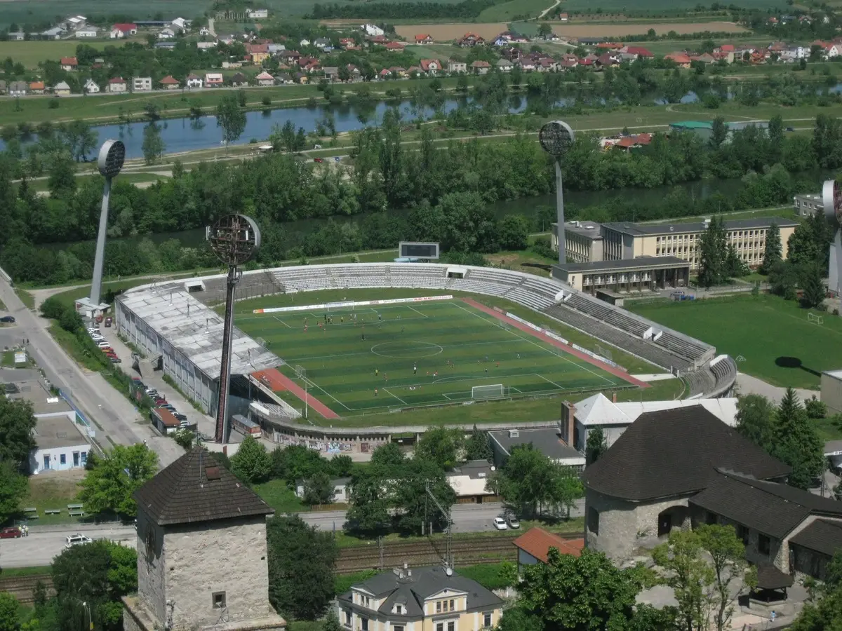 Vista aérea de un estadio de fútbol de tribunas bajas rodeado de arboleda y un río