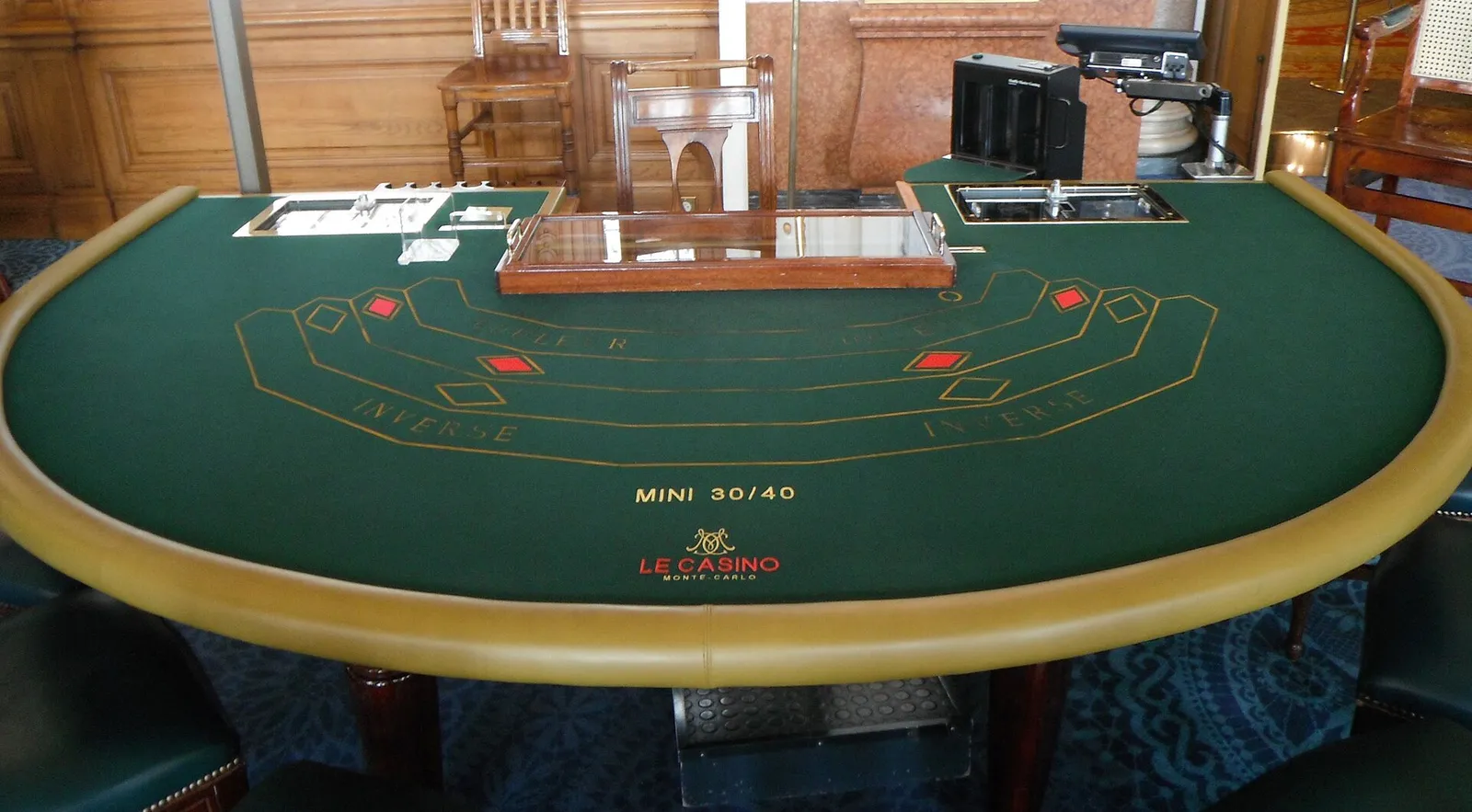 Mesa de casino de tapete verde vista desde el lado del jugador, con canto de madera clara y bandeja del crupier al fondo