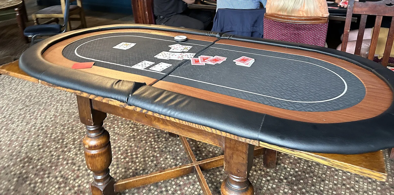 Mesa de póker ovalada con base de madera torneada, reposabrazos negro y cartas repartidas sobre el paño