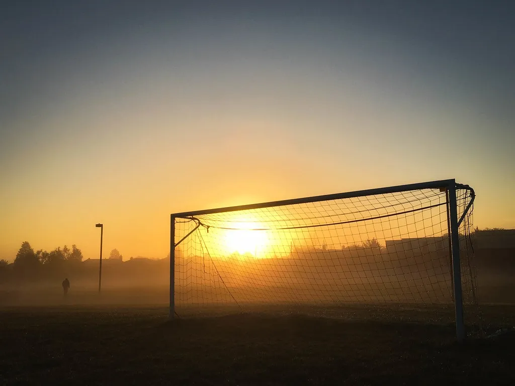 Portería de fútbol con red vieja recortada contra el amanecer sobre un campo con niebla baja